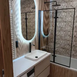 Nowoczesna łazienka z oświetlonym lustrem, kabiną prysznicową w czarnej ramie i umywalką nablatową, wykończona drewnianymi panelami i wzorzystymi płytkami.