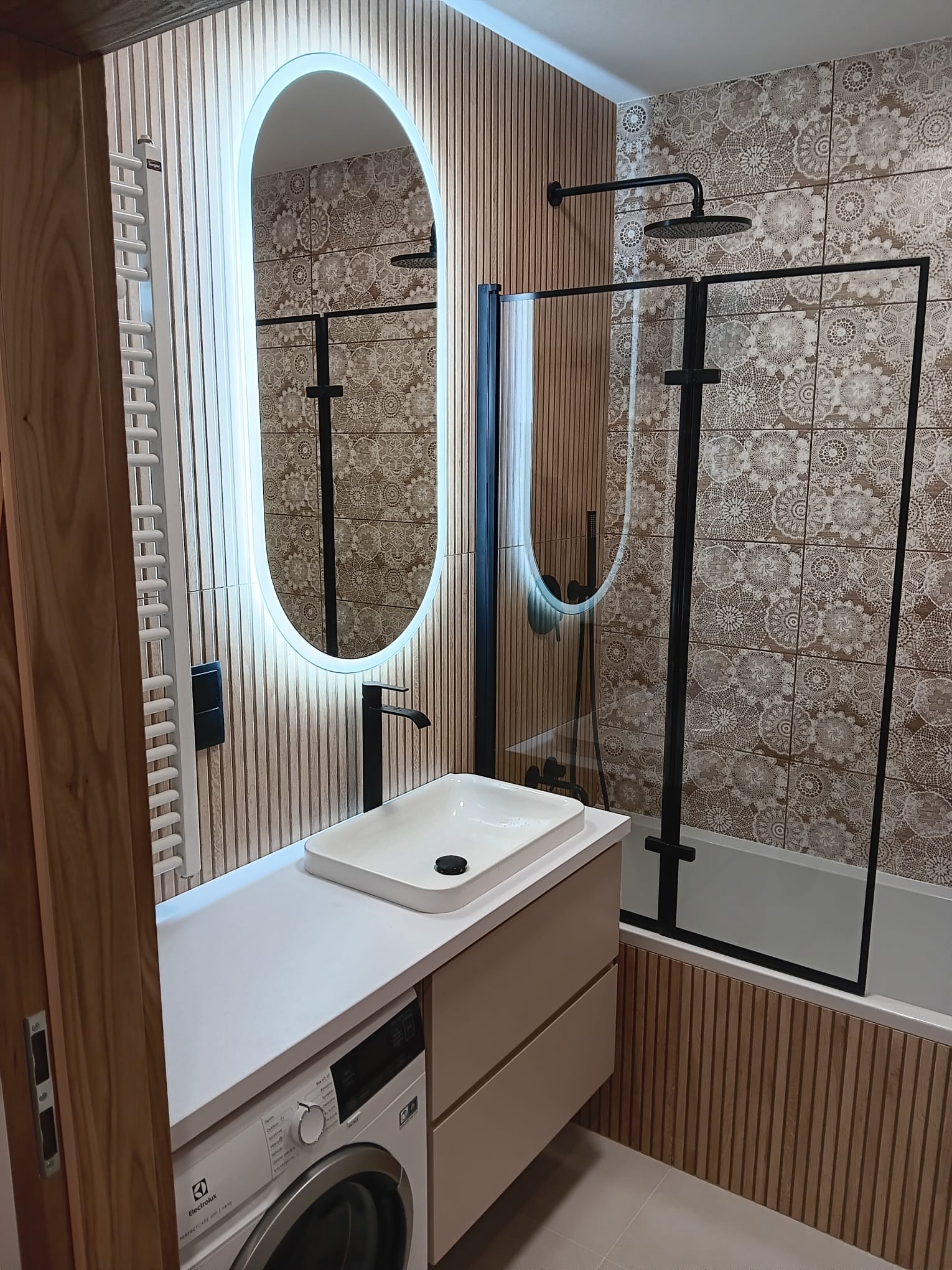 Nowoczesna łazienka z oświetlonym lustrem, kabiną prysznicową w czarnej ramie i umywalką nablatową, wykończona drewnianymi panelami i wzorzystymi płytkami.