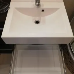 Biała, prostokątna umywalka ceramiczna z chromowaną baterią i otwartą szufladą szafki pod umywalką, widok z g&oacute;ry.