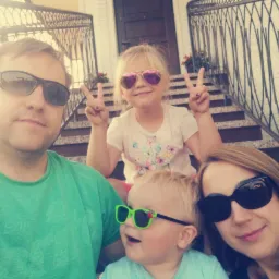 Rodzinne selfie na schodach przed domem z kutą balustradą. Dzieci w okularach pokazują znak zwycięstwa. Kwiaty w donicach na schodach. Jasne, słoneczne oświetlenie.