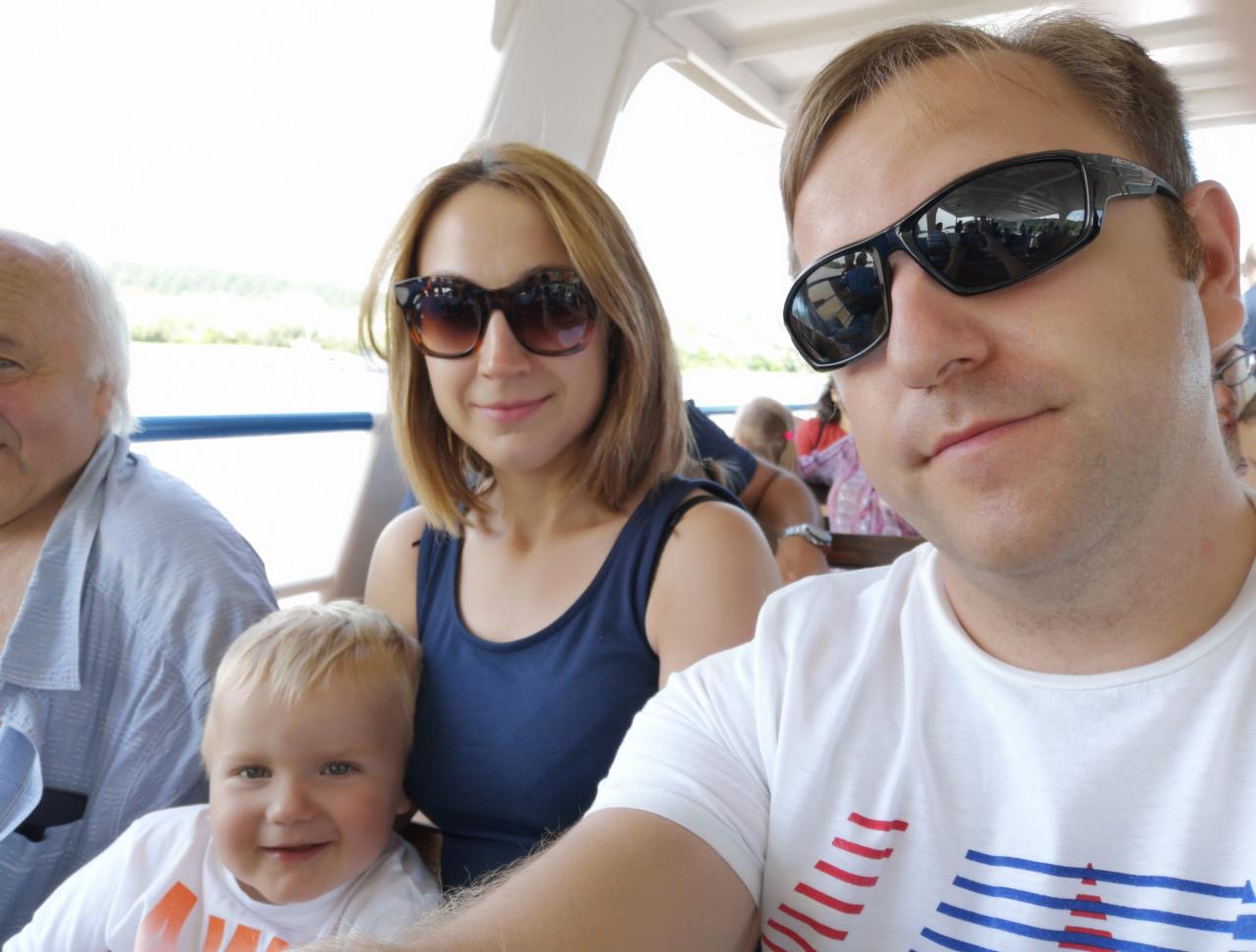 Rodzinne selfie na tle jeziora z łodzi. Uśmiechnięty blond chłopiec na pierwszym planie, za nim kobieta w okularach i mężczyzna z przodu, również w okularach.