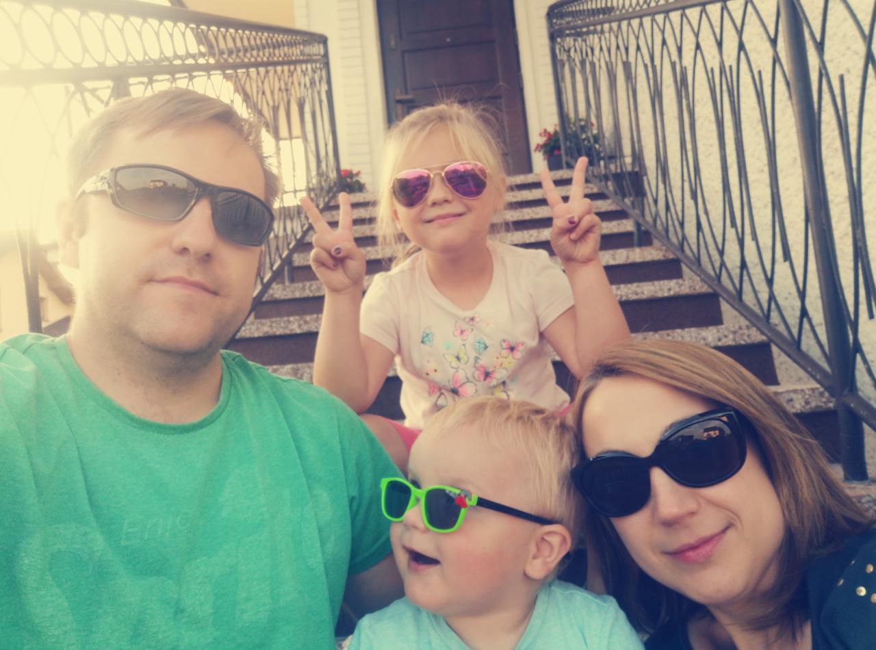 Rodzinne selfie na schodach przed domem z kutą balustradą. Dzieci w okularach pokazują znak zwycięstwa. Kwiaty w donicach na schodach. Jasne, słoneczne oświetlenie.