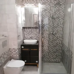 Nowoczesna łazienka z kabiną prysznicową typu walk-in, wiszącą toaletą i umywalką nablatową na ciemnej szafce, wykończona szaro-białą mozaiką.