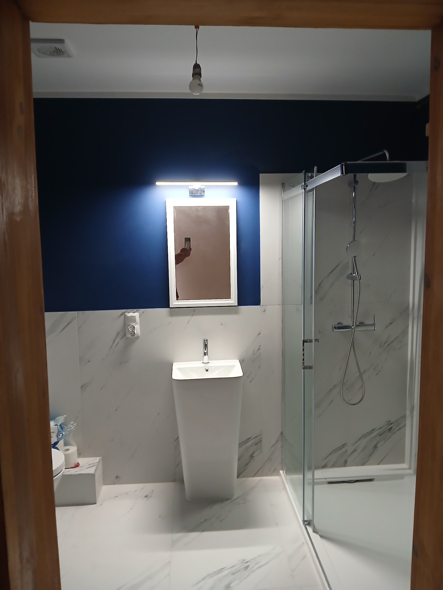 Wykończona łazienka z kabiną prysznicową, umywalką na postumencie i lustrem z oświetleniem. Ściany wyłożone płytkami imitującymi marmur, granatowa ściana u góry.