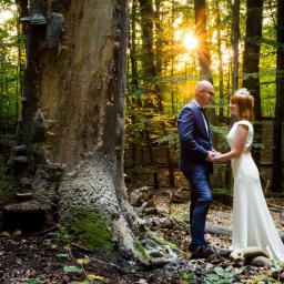 Foto Studio - Para trzymająca się za ręce w leśnej scenerii, mężczyzna w granatowym garniturze, kobieta w białej sukni, promienie słoneczne przebijają przez drzewa.