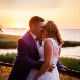 Foto Studio - Para młoda w objęciach na tle zachodzącego słońca nad jeziorem, mężczyzna w granatowym garniturze z muchą, kobieta w białej koronkowej sukni ślubnej.