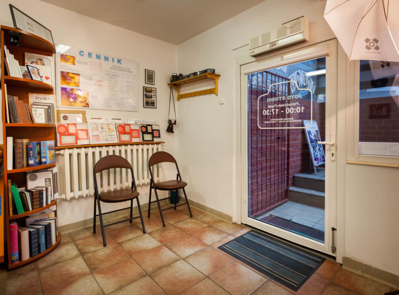 Wnętrze studia fotograficznego z cennikiem na ścianie, regałem z książkami i zdjęciami, dwoma krzesłami i widokiem na wejście przez oszklone drzwi.