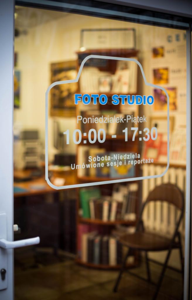 Witraż na szklanych drzwiach studia fotograficznego z godzinami otwarcia i informacją o sesjach i reportażach, widok na wnętrze z regałem pełnym książek i krzesłem.