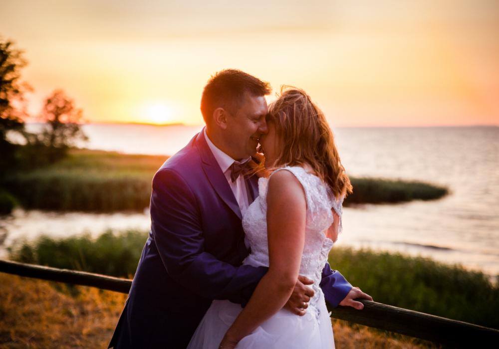 Para młoda w objęciach na tle zachodzącego słońca nad jeziorem, mężczyzna w granatowym garniturze z muchą, kobieta w białej koronkowej sukni ślubnej.