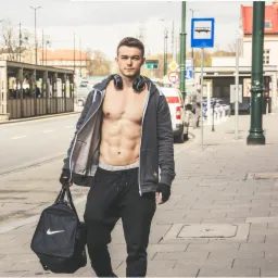 Młody mężczyzna z odsłoniętym torsem, w bluzie z kapturem i słuchawkami na szyi, trzymający torbę sportową Nike na tle miejskiego krajobrazu z przystankiem autobusowym.