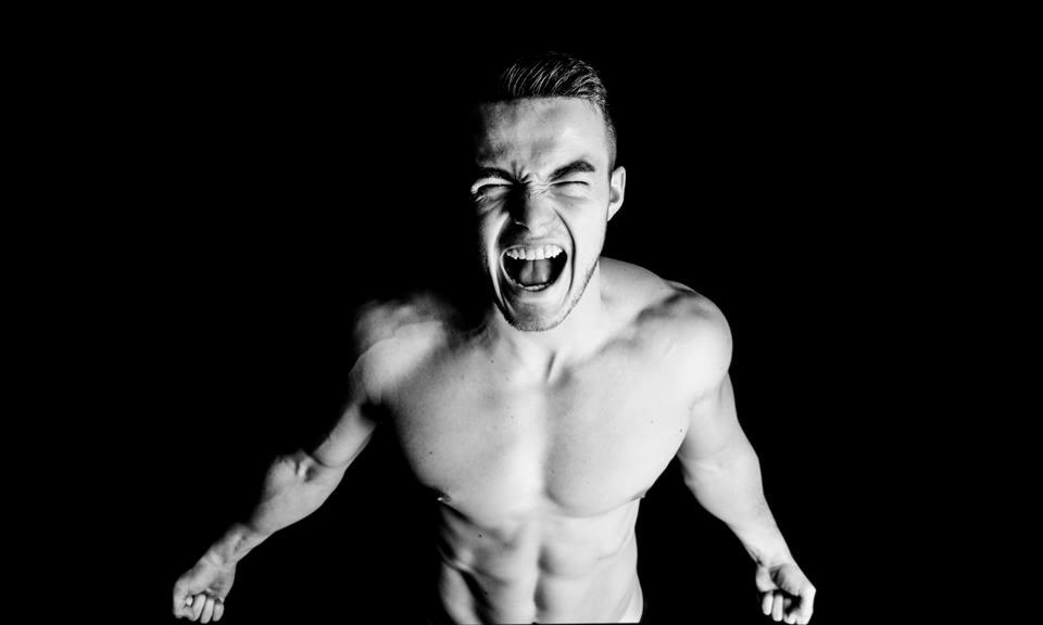 Czarno-białe zdjęcie muskularnego mężczyzny z szeroko otwartymi ustami i zaciśniętymi oczami, oświetlonego od dołu, na czarnym tle.