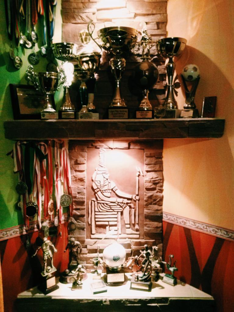 Kolekcja trofeów, medali i statuetek sportowych na półce i komodzie z ceglanym tłem, oświetlona ciepłym światłem.