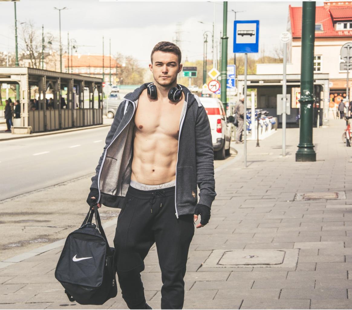 Młody mężczyzna z odsłoniętym torsem, w bluzie z kapturem i słuchawkami na szyi, trzymający torbę sportową Nike na tle miejskiego krajobrazu z przystankiem autobusowym.