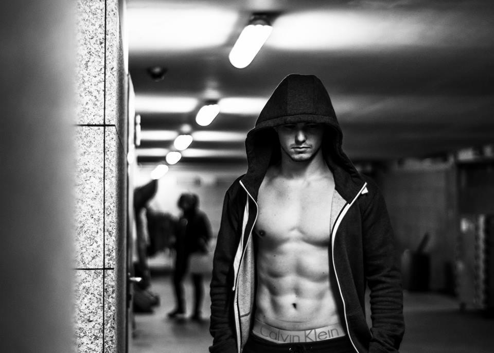 Czarno-białe zdjęcie mężczyzny z odsłoniętym torsem w bluzie z kapturem, stojącego w oświetlonym tunelu z widocznymi osobami w tle.