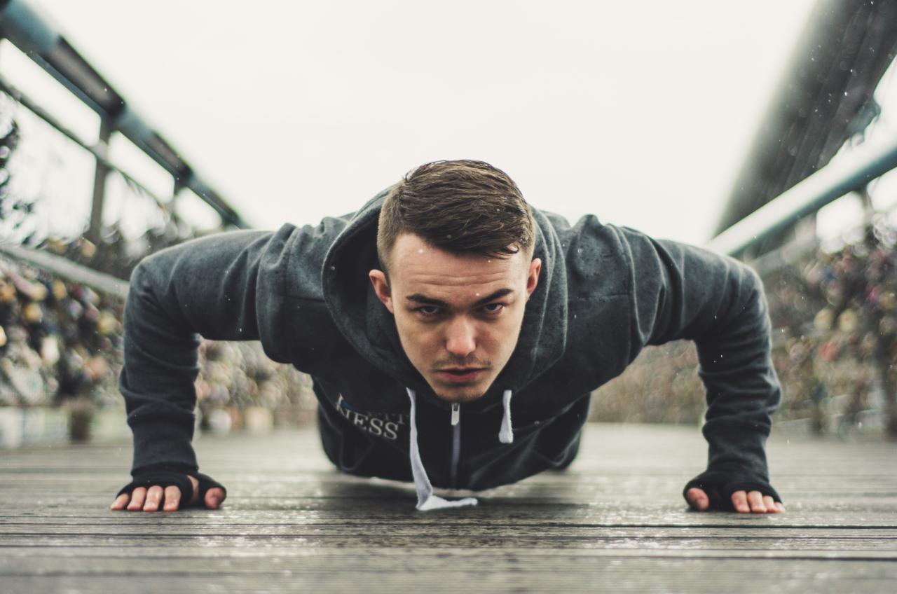 Młody mężczyzna w szarej bluzie z kapturem, robiący pompki na drewnianym pomoście w pochmurny dzień, widok z bliska.