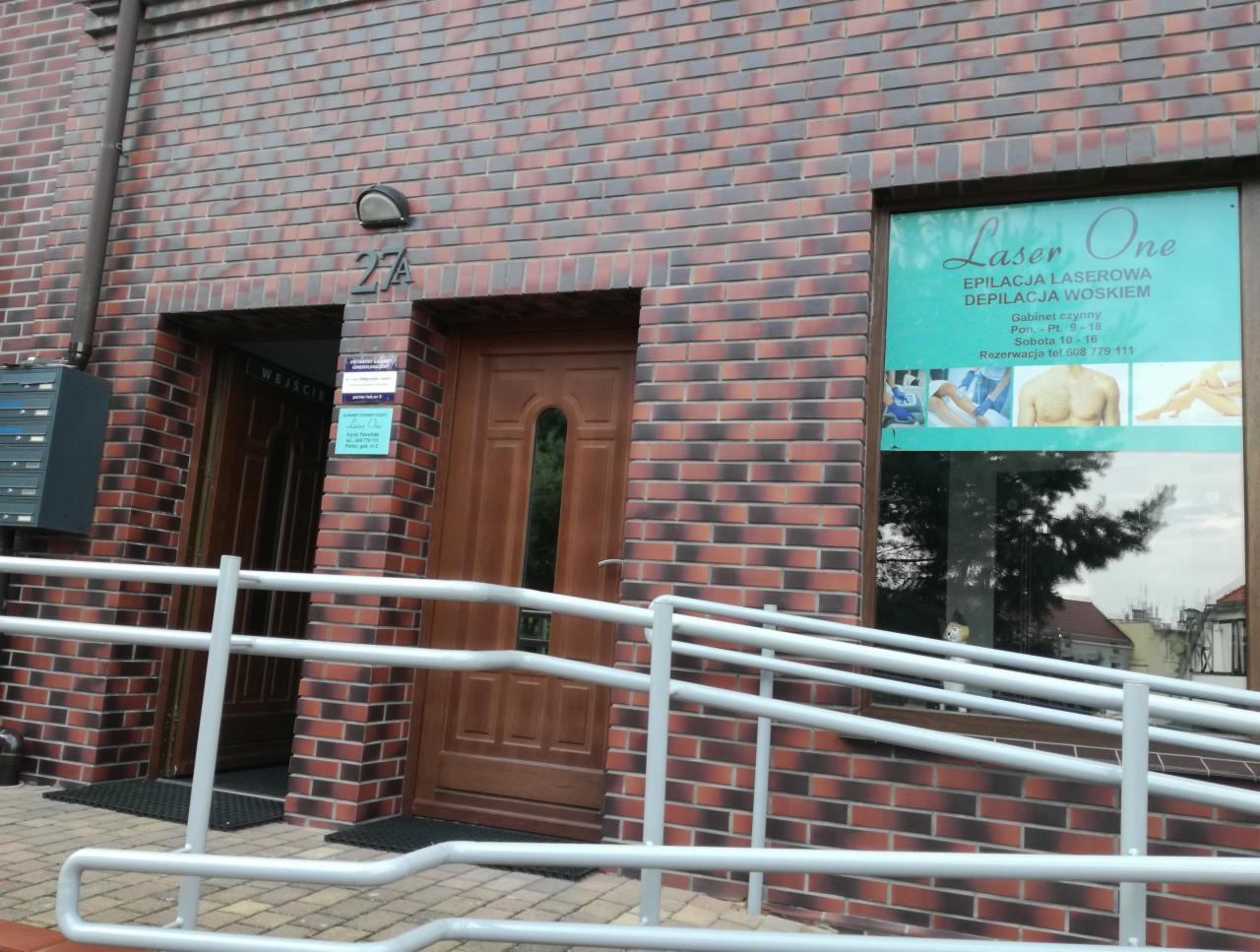 Wejście do budynku z cegły z szyldem 'Laser One' oferującego epilację laserową i depilację woskiem, widoczna rampa dla niepełnosprawnych.