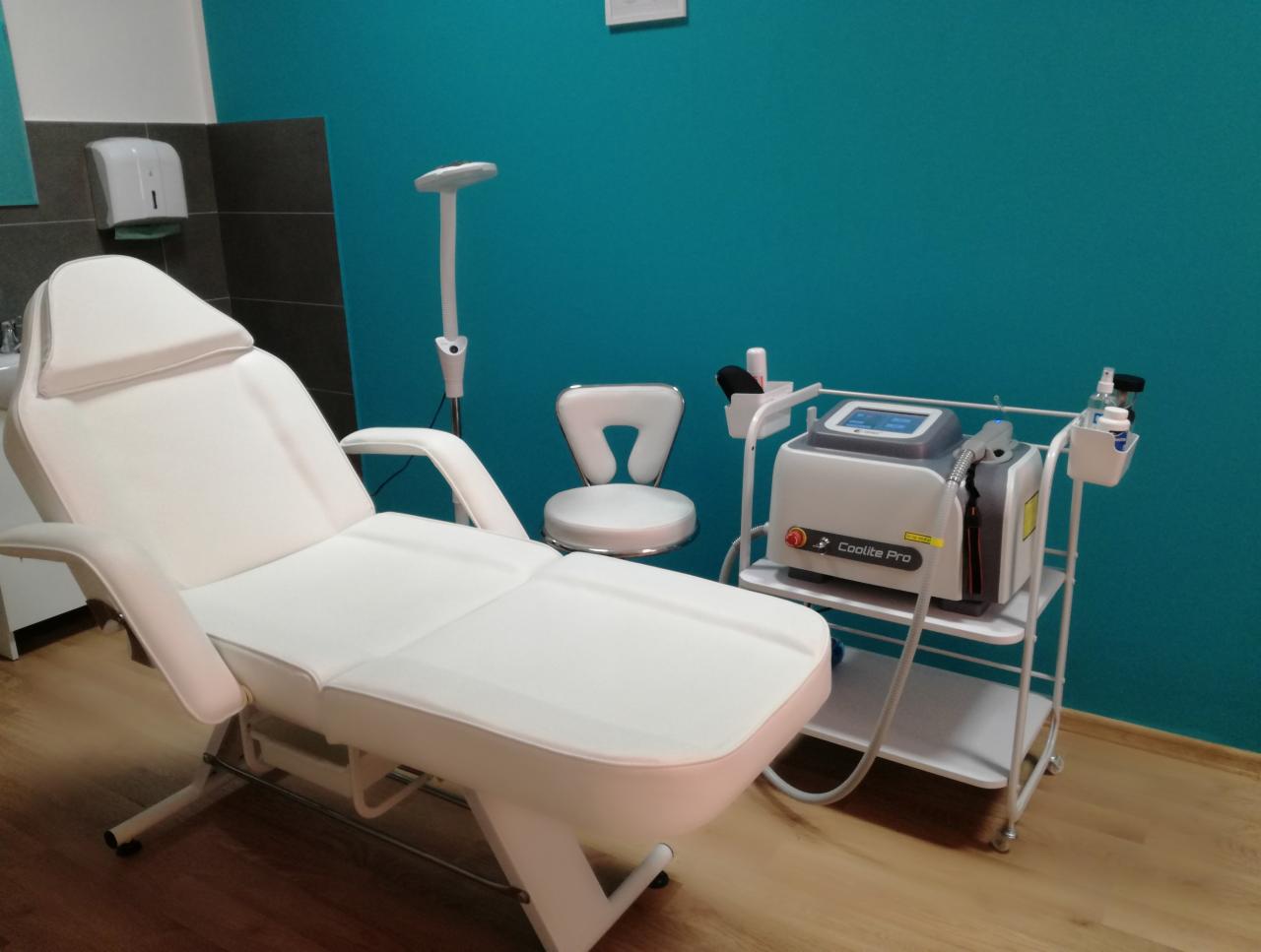 Jasne wnętrze gabinetu kosmetycznego z białym fotelem do zabiegów, lampą podłogową i urządzeniem Coolite Pro na wózku, na tle turkusowej ściany.