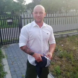 Mężczyzna w białej koszuli stoi na chodniku przed ogrodzeniem, trzymając w rękach granatową bluzę z białymi paskami; widoczny tatuaż na ramieniu.