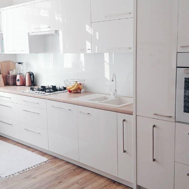 Jasna, minimalistyczna kuchnia z białymi, lakierowanymi frontami, drewnianym blatem, dwukomorowym zlewem i metalowym koszykiem z owocami.