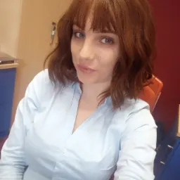 Portret kobiety z rudymi włosami i niebieskimi oczami, ubrana w jasnoniebieską koszulę, siedzącej na pomarańczowym krześle w biurze.