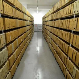 Magazyn z rzędami kartonowych pudeł archiwizacyjnych, oznaczonych napisami, ustawionych na metalowych regałach, widok z perspektywy podłogi.