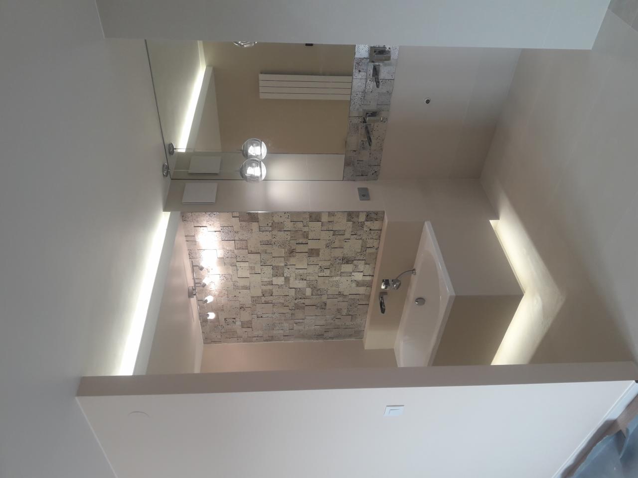 Nowoczesna łazienka po remoncie: beżowe ściany, umywalka z baterią, lustro z oświetleniem i dekoracyjna ściana z mozaiki. Widok na sufit z oświetleniem LED.