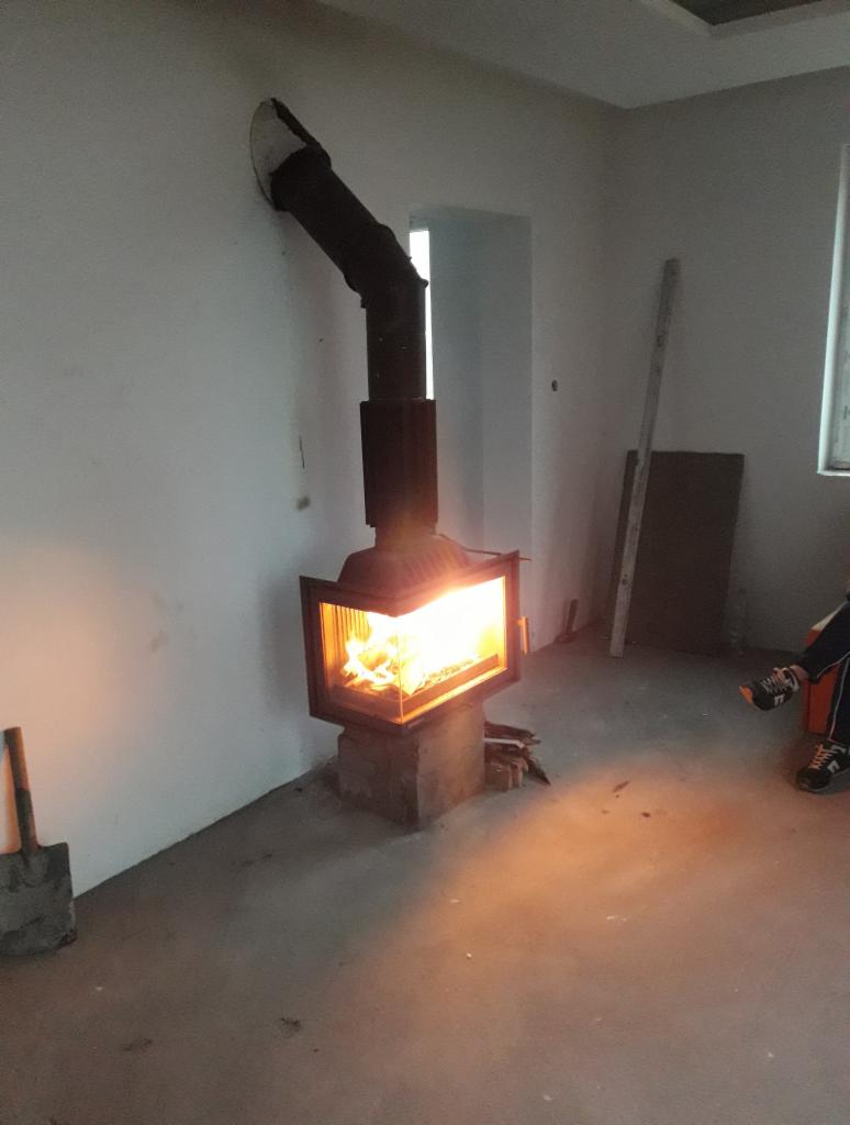 Nowo zainstalowany kominek z czarnym przewodem kominowym w surowym wnętrzu, z widoczną łopatą i fragmentem osoby w prawym dolnym rogu.