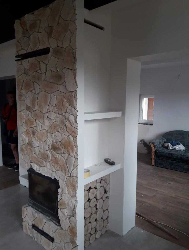 Wykończenie ściany z kamienia dekoracyjnego z wbudowanym kominkiem i półkami z dekoracją z drewna, widoczne niedoskonałości wykończenia.
