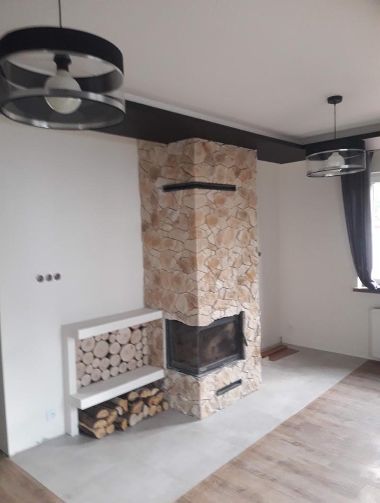 Kominek obłożony kamieniem z wkładem kominkowym, z półką na drewno, nowoczesne lampy wiszące w minimalistycznym wnętrzu.