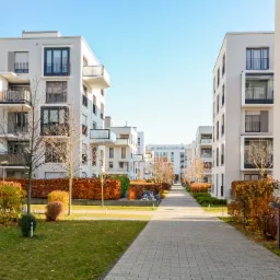 Nowoczesne osiedle apartamentowców z białymi elewacjami, balkonami z czarnymi balustradami, widok z perspektywy alei spacerowej z kostki brukowej, uporządkowana zieleń.