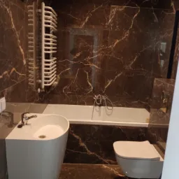 Wykończona łazienka z brązowymi marmurkowymi płytkami, białą umywalką nablatową, wanną, toaletą w zabudowie i grzejnikiem drabinkowym.