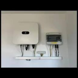 Biały falownik fotowoltaiczny marki Huawei i skrzynka elektryczna zamontowane na białej ścianie, połączone przewodami na białej listwie przypodłogowej z oznaczeniem PV.