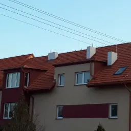 Budynek wielorodzinny z czerwoną dachówką, kominami i instalacją odgromową na dachu, widoczne przewody elektryczne nad budynkiem.