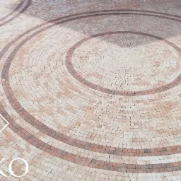 Okładzina z kostki brukowej ułożona w koncentryczne okręgi, widoczny fragment logo firmy na pierwszym planie.