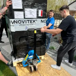 Dwóch mężczyzn transportuje za pomocą wózka paletowego dużą, owiniętą folią paczkę marki Novitek na palecie, po drewnianej rampie na zewnątrz budynku.