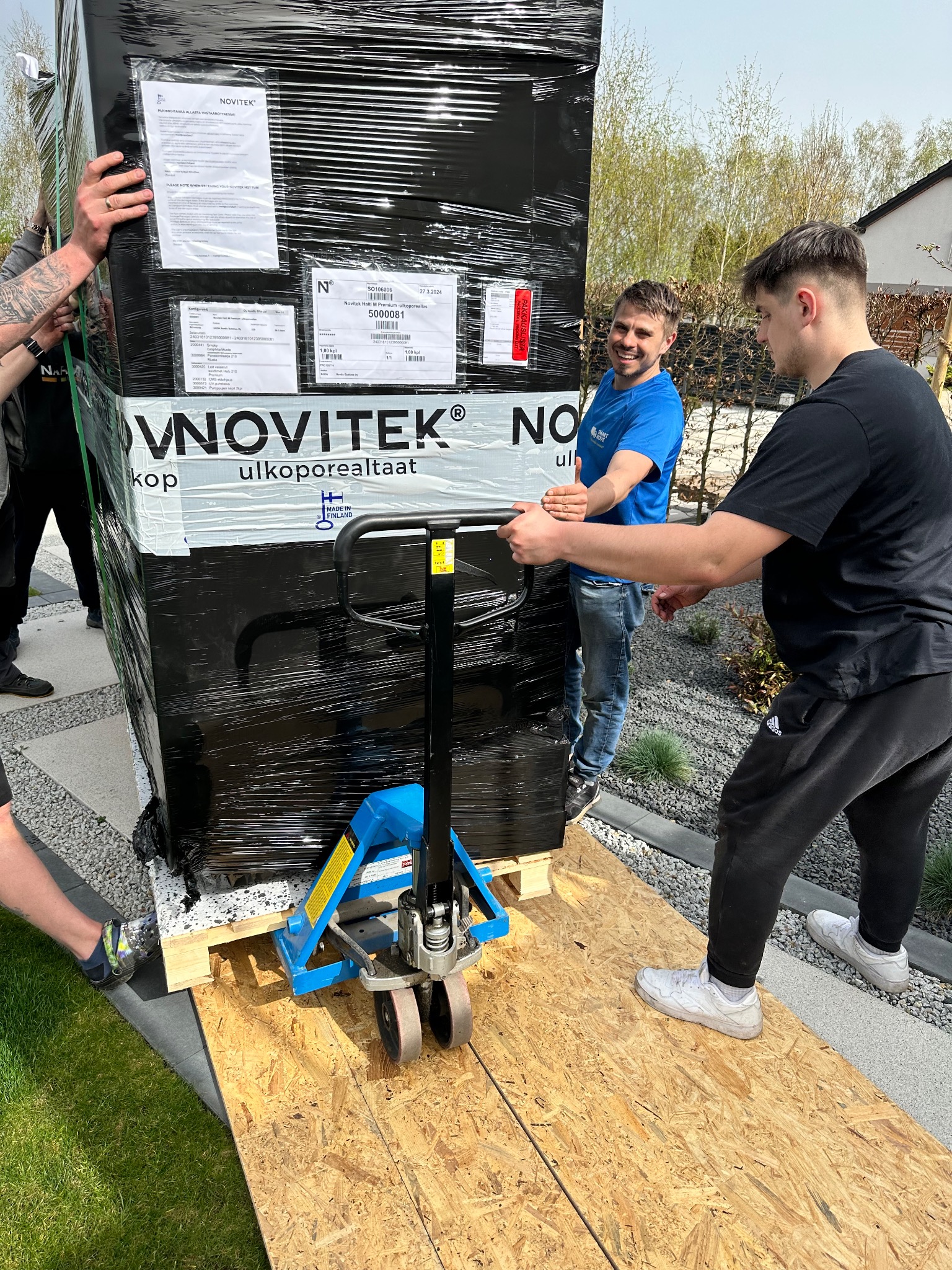 Dwóch mężczyzn transportuje za pomocą wózka paletowego dużą, owiniętą folią paczkę marki Novitek na palecie, po drewnianej rampie na zewnątrz budynku.