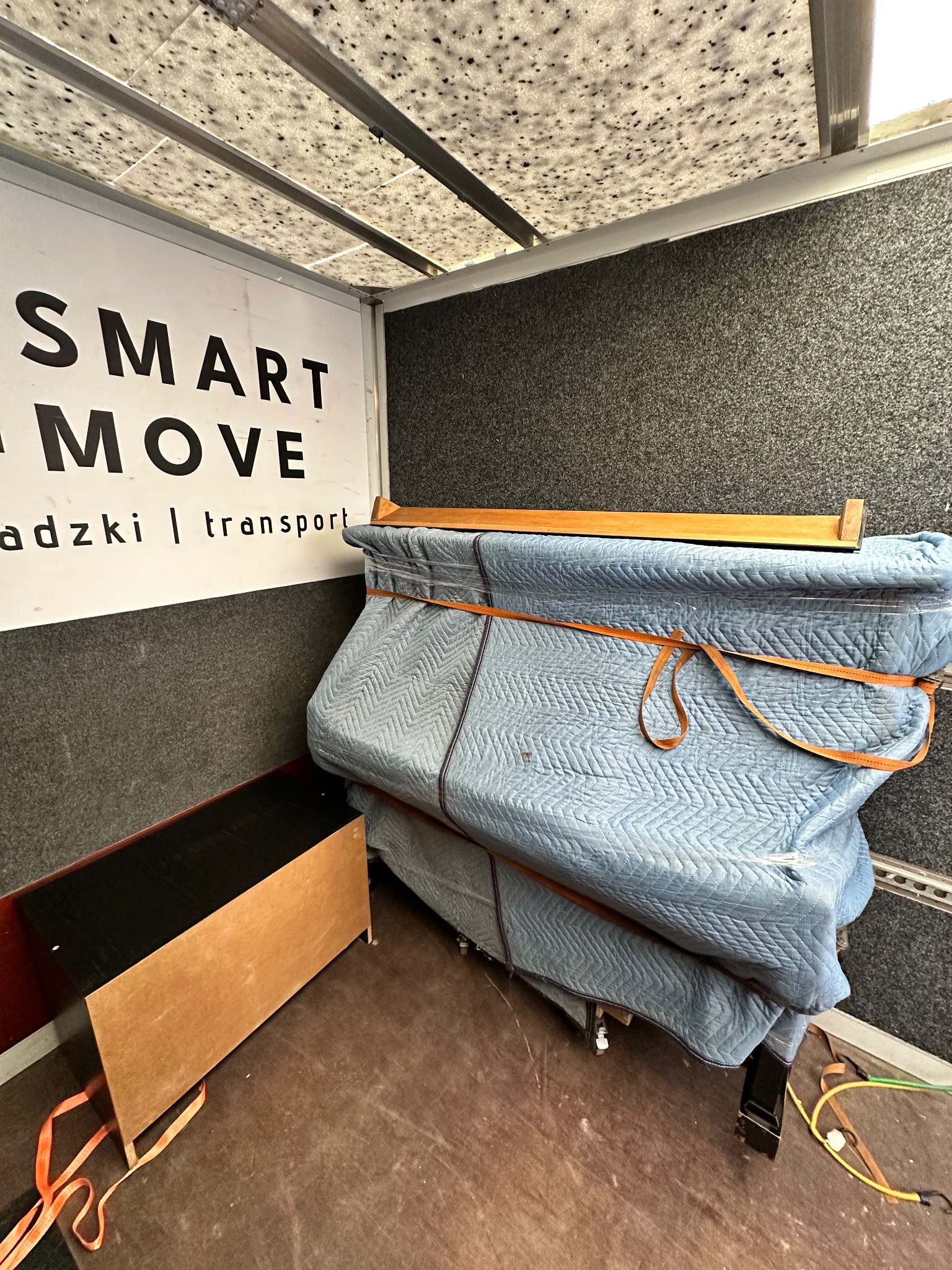 Wnętrze furgonetki z zabezpieczonym meblem owiniętym w pikowaną, błękitną folię i spiętym pomarańczowymi pasami, obok stoi ciemny mebel, na ścianie widoczne logo firmy transportowej.