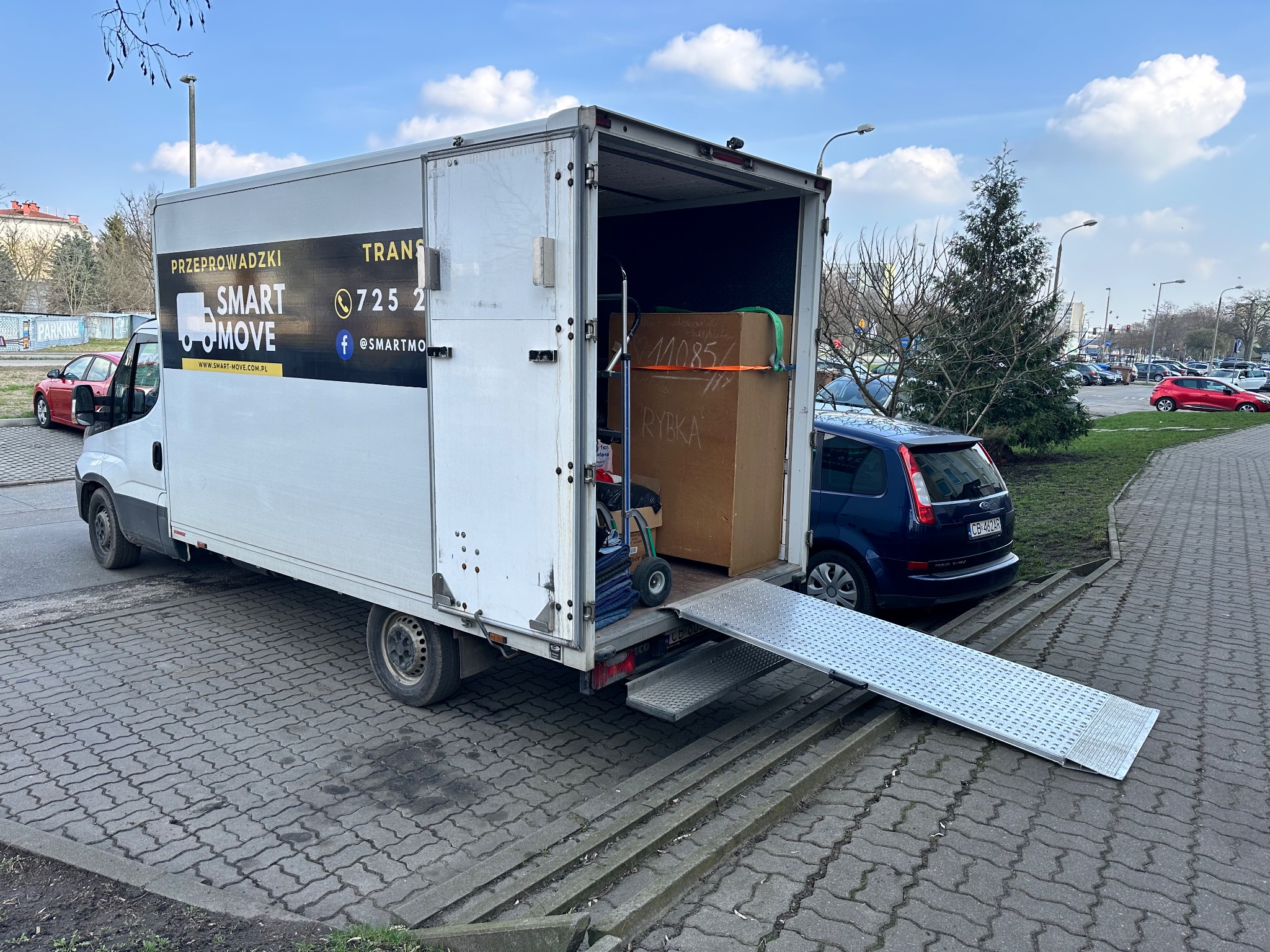 Samochód dostawczy firmy przeprowadzkowej Smart Move z otwartą rampą załadunkową, częściowo załadowany kartonami, zaparkowany na brukowanym parkingu. Na boku pojazdu widoczne logo i dane kontaktowe...