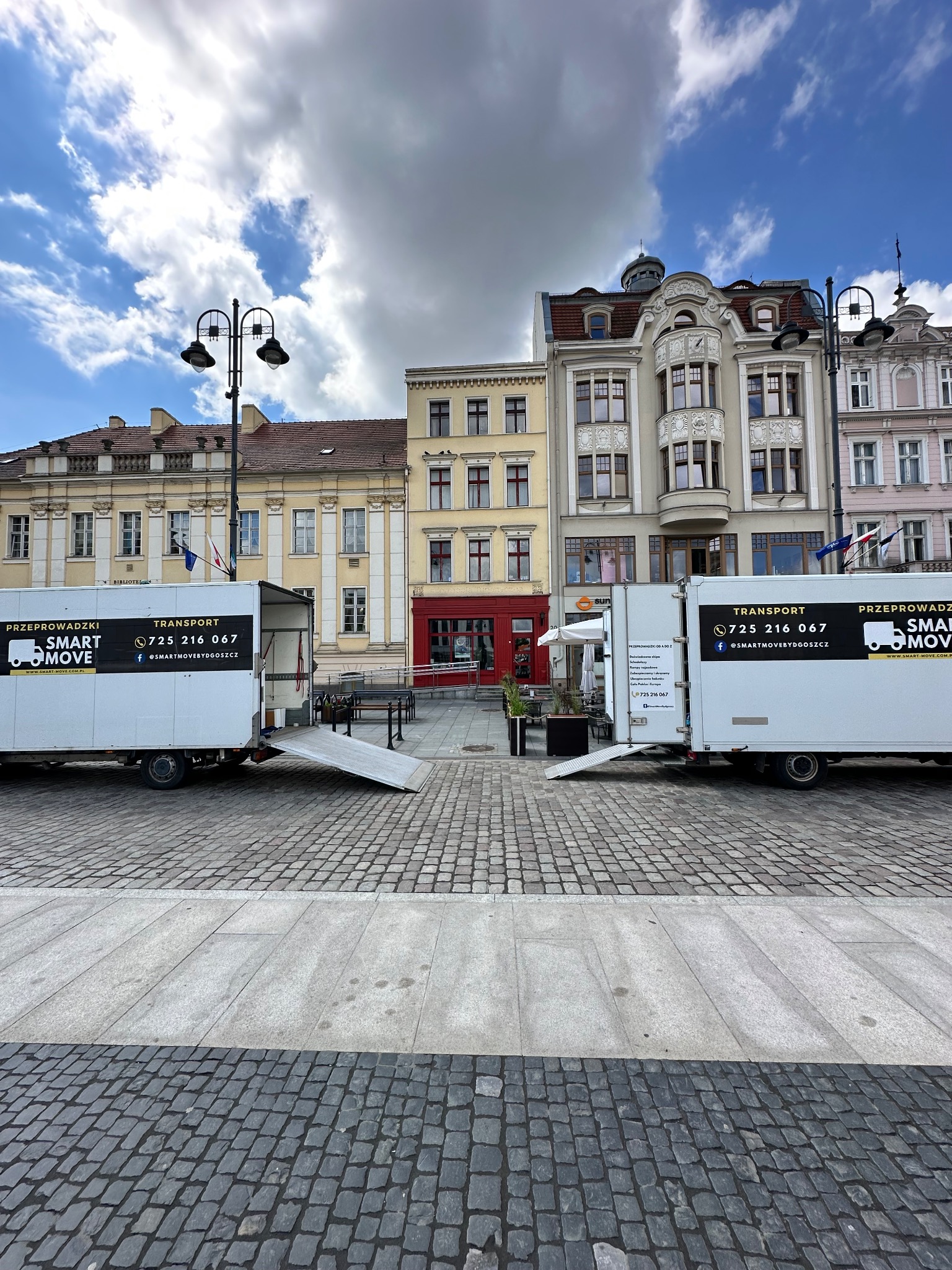 Dwa białe samochody dostawcze firmy przeprowadzkowej Smart Move z otwartymi rampami na brukowanym placu miejskim w Bydgoszczy, na tle kolorowych kamienic i błękitnego nieba z chmurami.