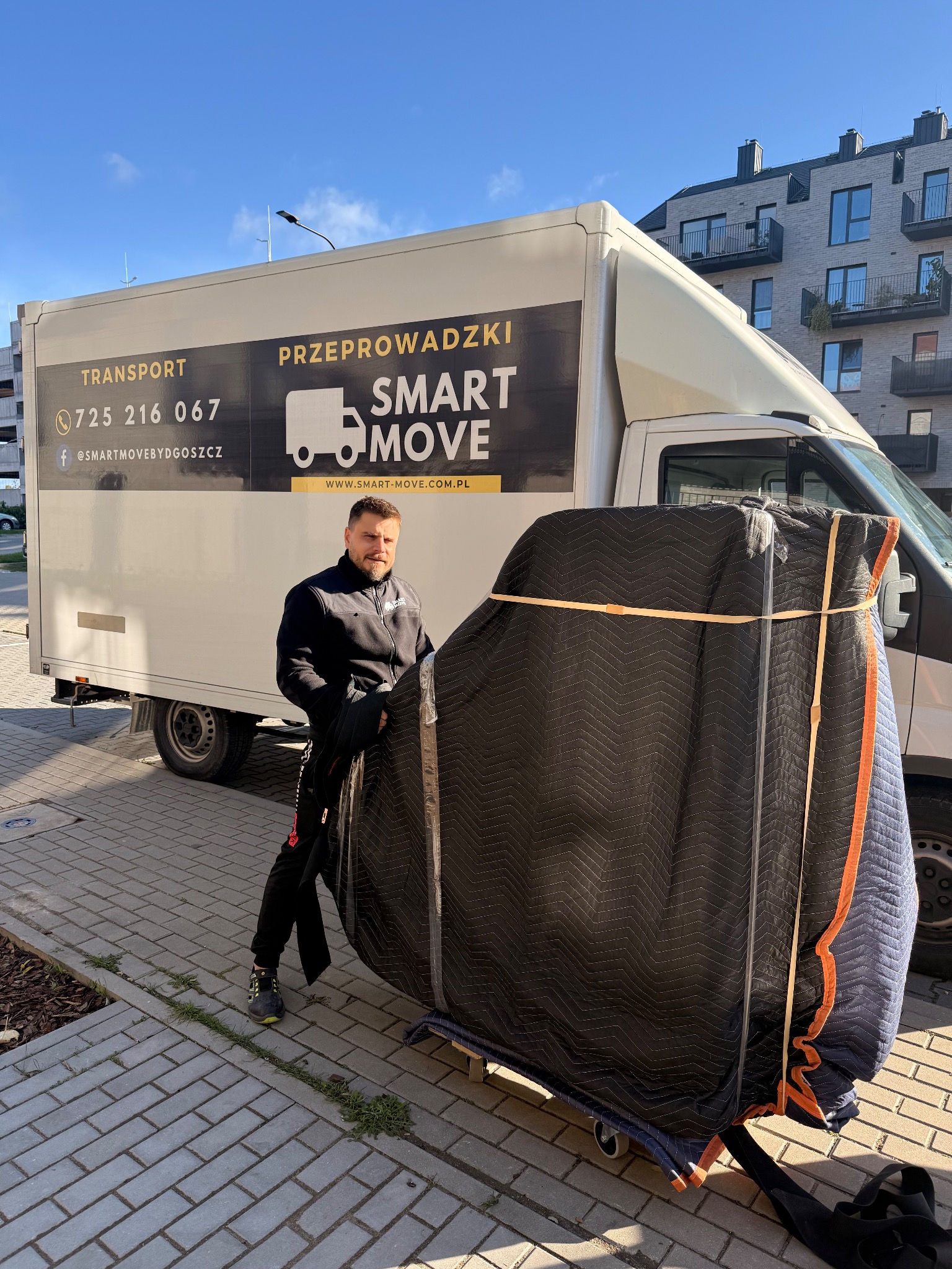 Mężczyzna przy samochodzie dostawczym z logo firmy przeprowadzkowej, zabezpieczony mebel na wózku, budynek mieszkalny w tle, słoneczny dzień.