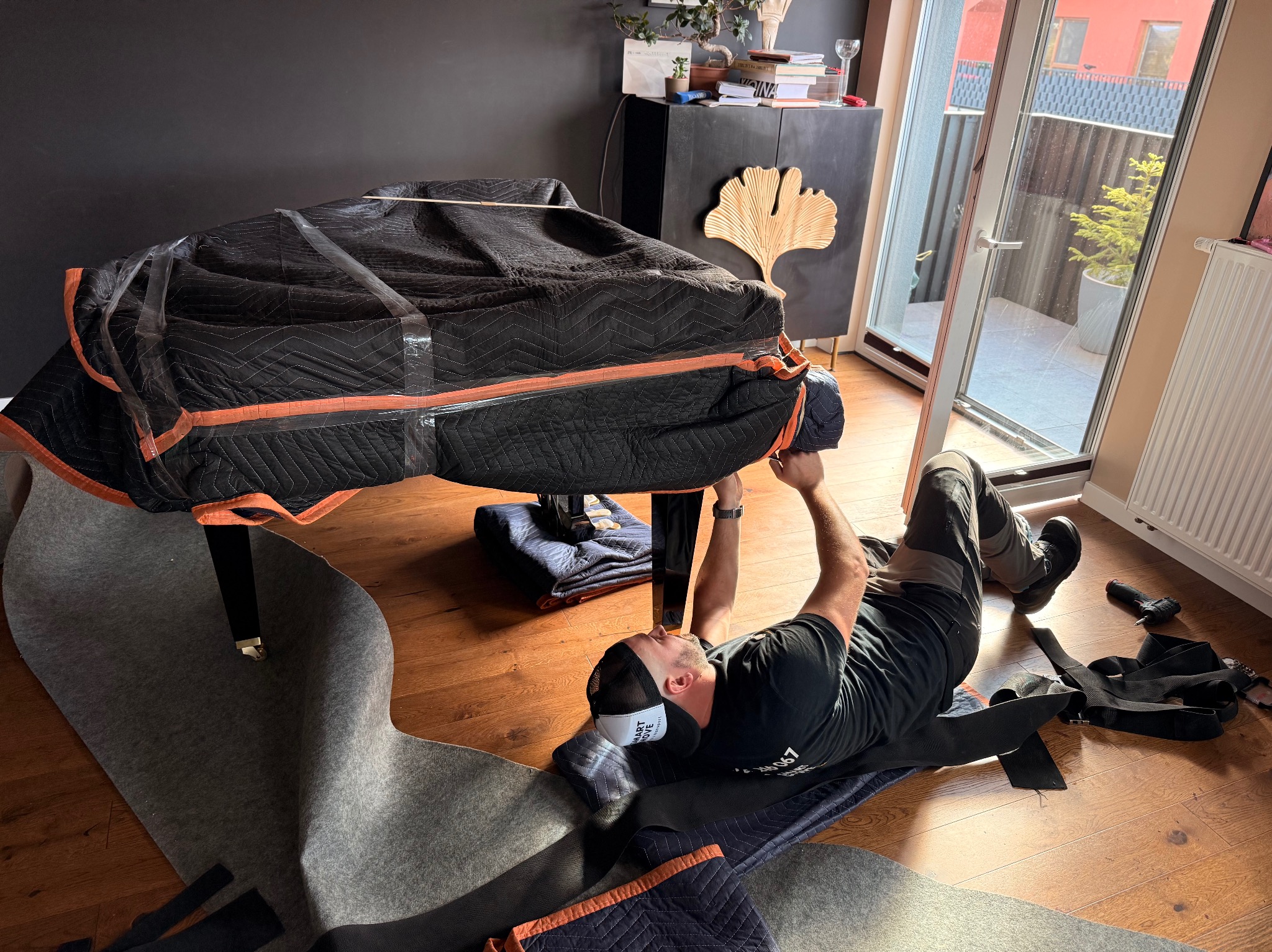 Pracownik firmy przeprowadzkowej zabezpiecza pianino do transportu. Pianino owinięte w koc transportowy, podłoga zabezpieczona matą. Widoczne pasy transportowe i narzędzia.