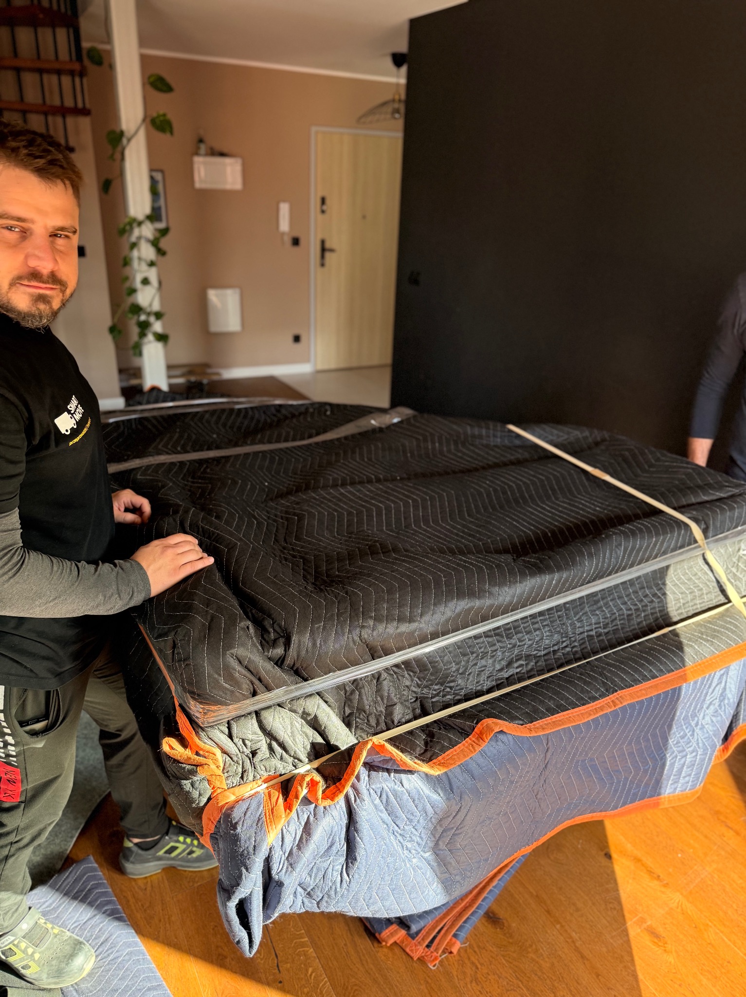 Pracownik firmy przeprowadzkowej zabezpiecza pianino kocami transportowymi i folią stretch przed przeniesieniem. Widoczny fragment wnętrza mieszkania.