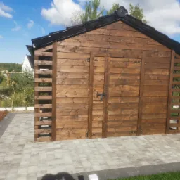 Drewniana altana ogrodowa z czarnym dachem, widok z przodu na tle błękitnego nieba i zieleni, wykończona kostką brukową.