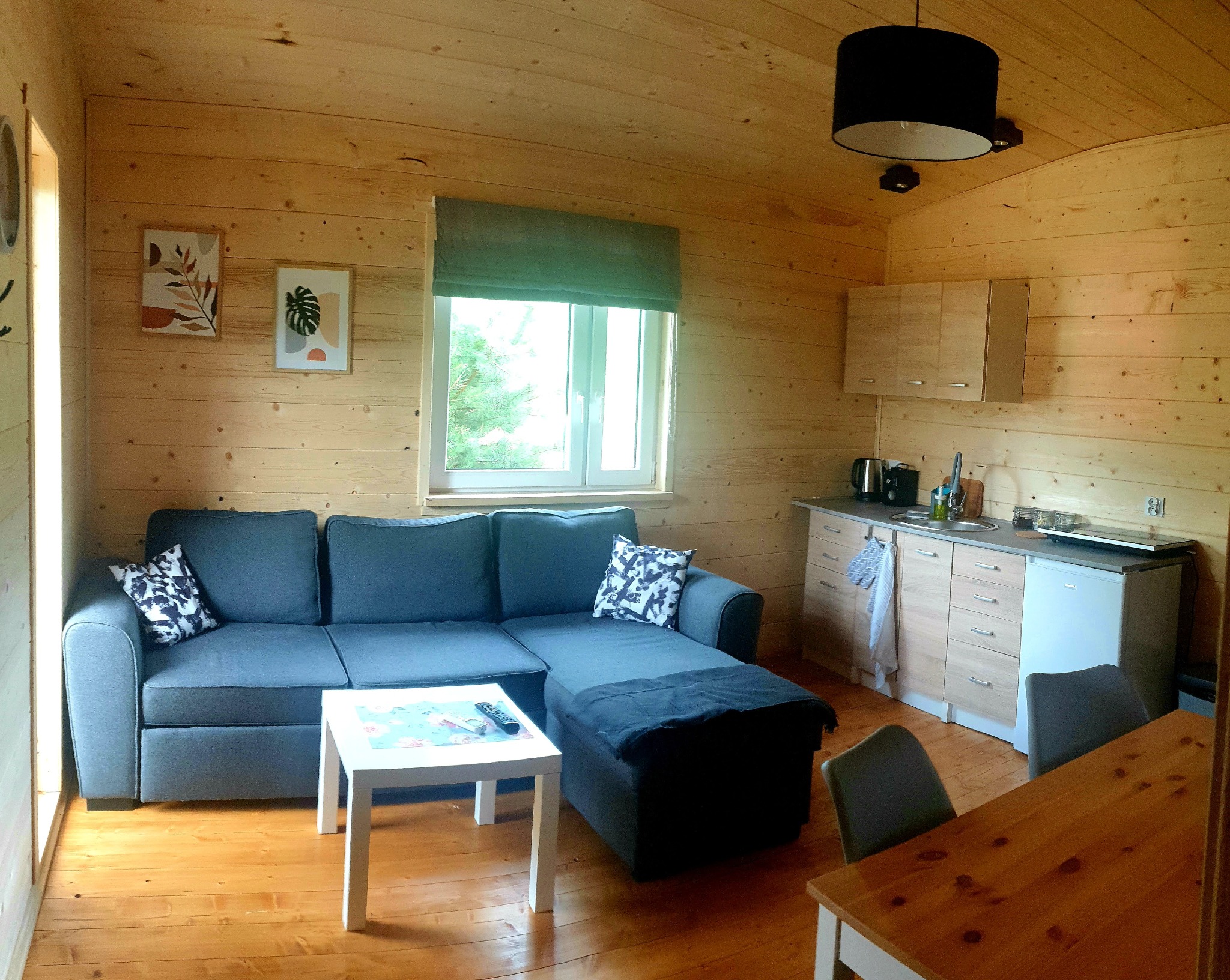 Wnętrze drewnianego domu z szarą sofą narożną, białym stolikiem kawowym, aneksem kuchennym i drewnianym stołem jadalnym, oświetlone czarną lampą wiszącą.