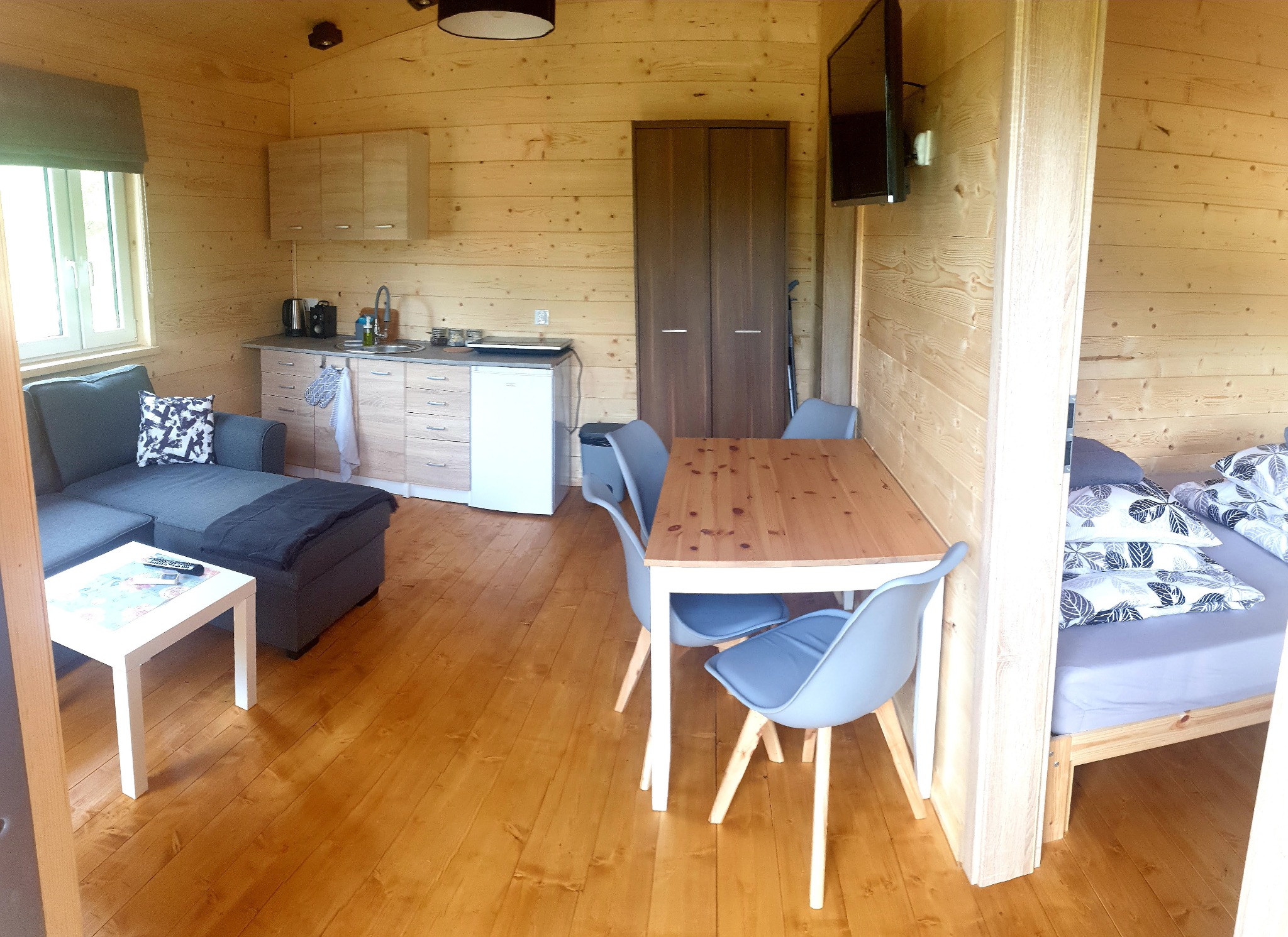 Wnętrze małego domku z drewnianych bali z aneksem kuchennym, częścią wypoczynkową z szarą sofą i stolikiem oraz wydzieloną sypialnią z łóżkiem.