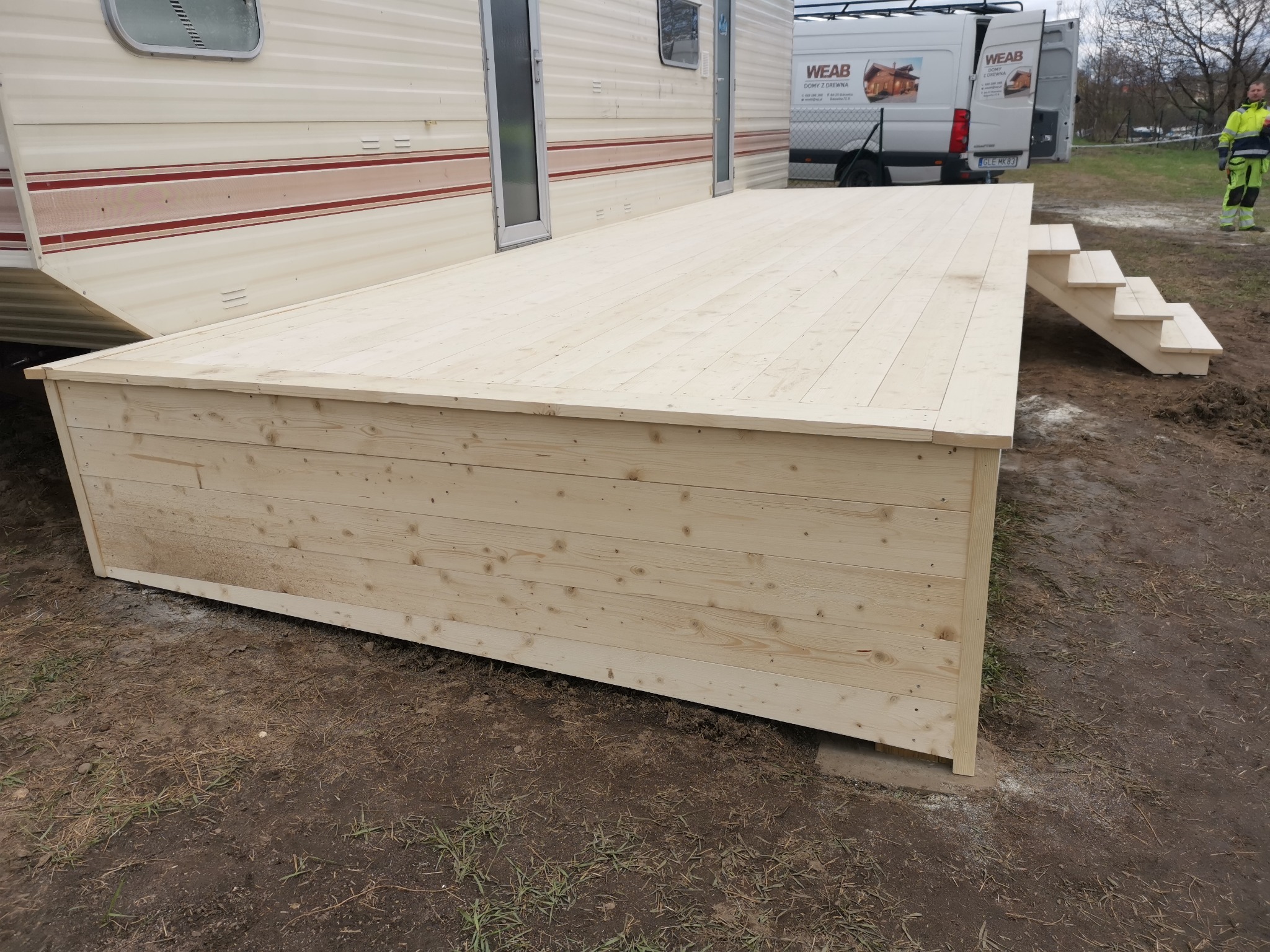 Drewniany taras z jasnego drewna z niskimi schodkami, przylegający do przyczepy kempingowej, na ziemi z widocznym samochodem dostawczym w tle.