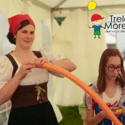 Kobieta w stroju pirata modeluje pomarańczowy balon na długi kształt, obok druga kobieta w stroju Królewny Śnieżki, w tle widoczne logo firmy Trele Morele.