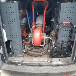 Wnętrze samochodu serwisowego z widoczną maszyną do czyszczenia kanalizacji z czerwoną obudową i spiralnym wężem, obok regały z narzędziami i akcesoriami hydraulicznymi.