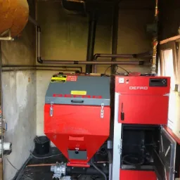 Kotłownia z kotłem na paliwo stałe marki Defro, z widocznymi rurami instalacji grzewczej, zbiornikiem wyrównawczym i elementami instalacji wodnej w pomieszczeniu o surowych, betonowych ścianach.