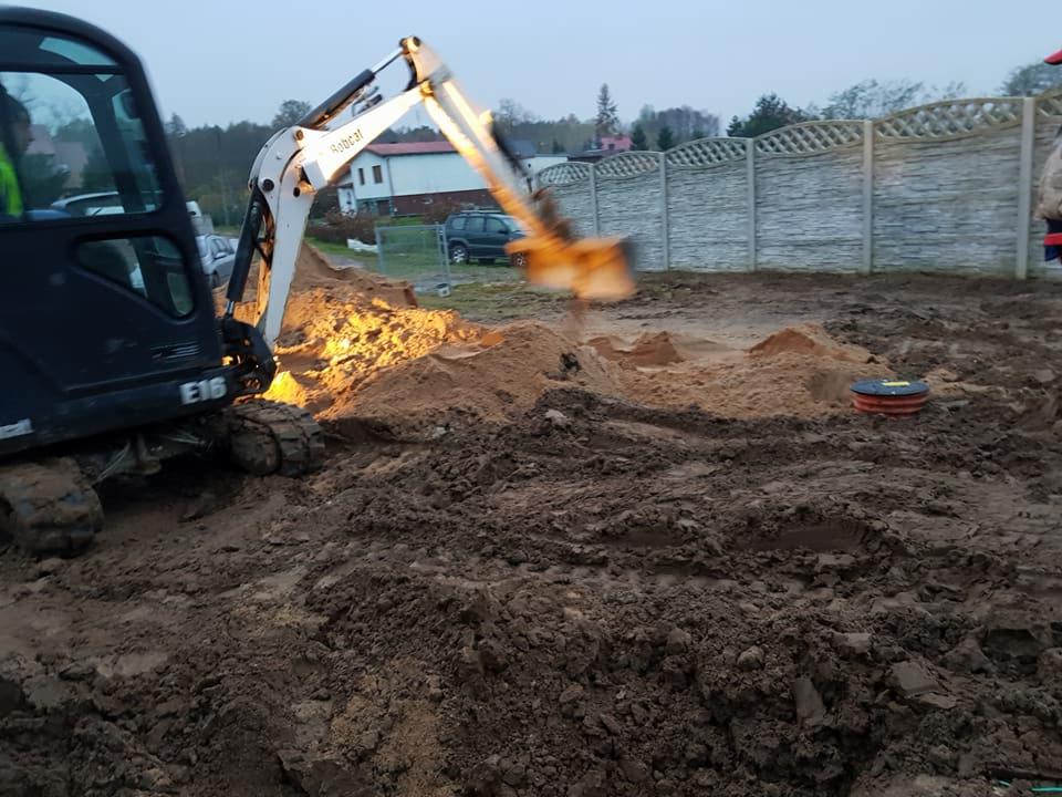 Mini koparka Bobcat E16 podczas prac ziemnych w ogrodzie, prawdopodobnie przygotowanie terenu pod instalację, widoczny fragment betonowego ogrodzenia i studzienka kanalizacyjna.